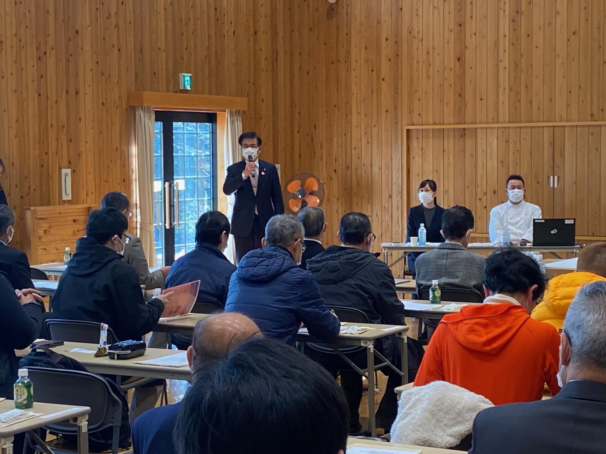 木目調の壁の集会所で、マスクを着けた市長がマイクを持って聴衆に話している様子を写した写真