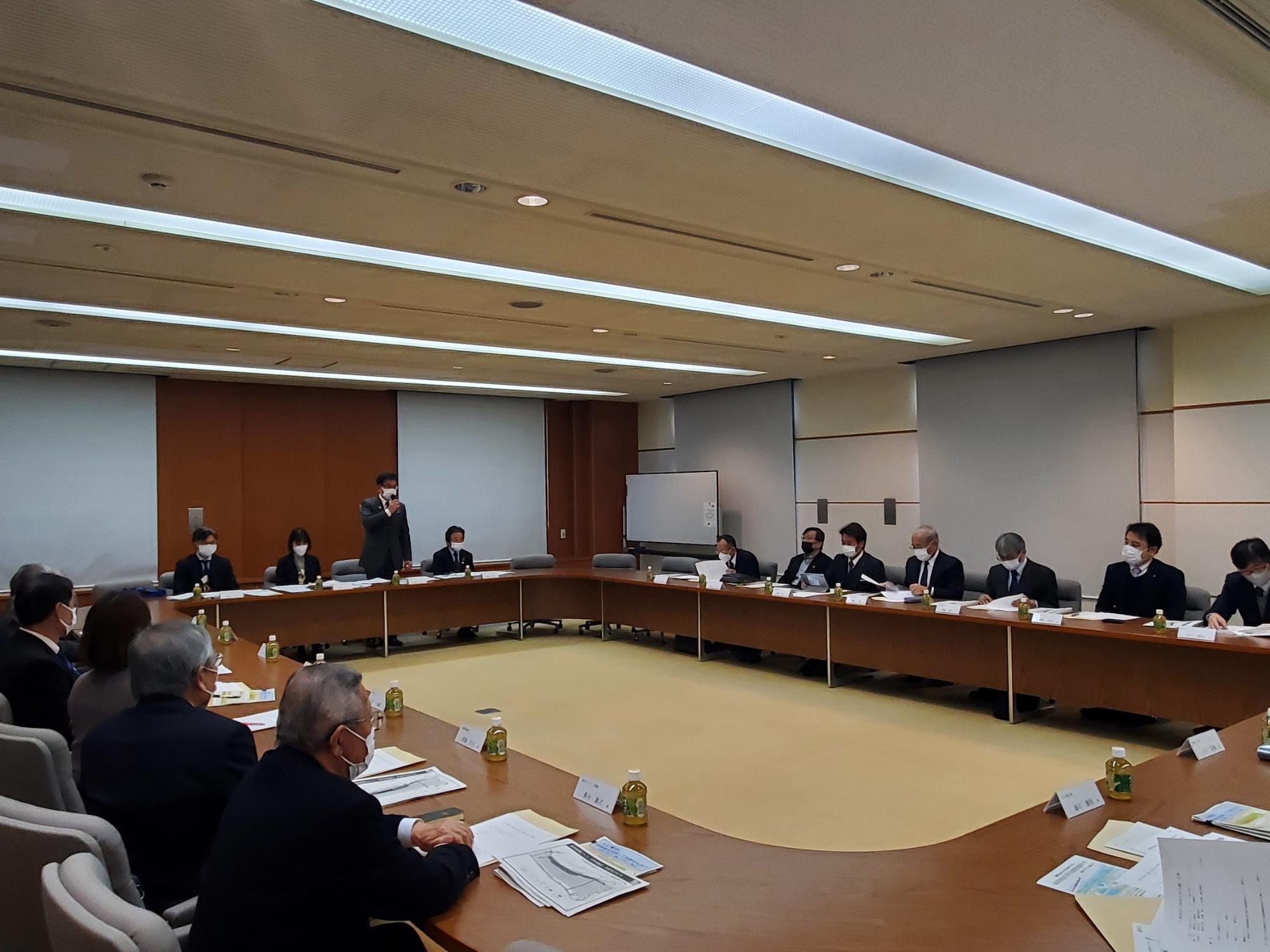 大会議室のコの字型に配置された大きなテーブルを囲んで、複数のスーツ姿の関係者が着席し、中央に立つ市長が発言している様子を捉えた会議風景の写真