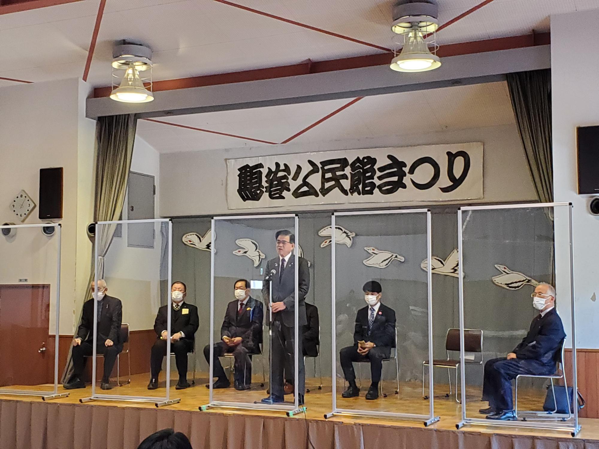 檀上の中央でスーツ姿の市長がマイクを持ってスピーチをし、その両側にアクリル板で仕切られた椅子に座る複数の男性たちがいる様子を捉えた写真