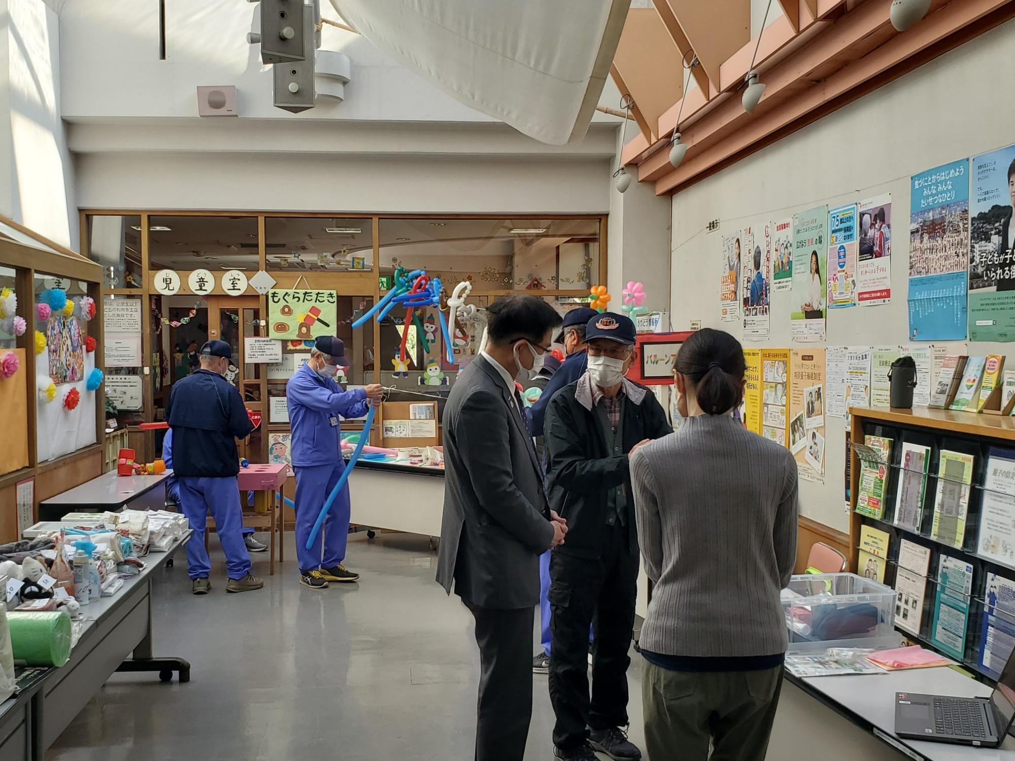 公民館のような室内で、市長と複数人が集まって談笑する様子や、風船アートを楽しむ人、そして壁に貼られた多数のポスターなどを捉えたイベント風景の写真