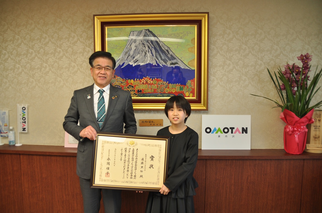 スーツを着た市長が、黒い服を着た遠藤里桜さんと共に額に入った表彰状を持ち、カメラに向かって微笑んでいる様子を捉えた写真