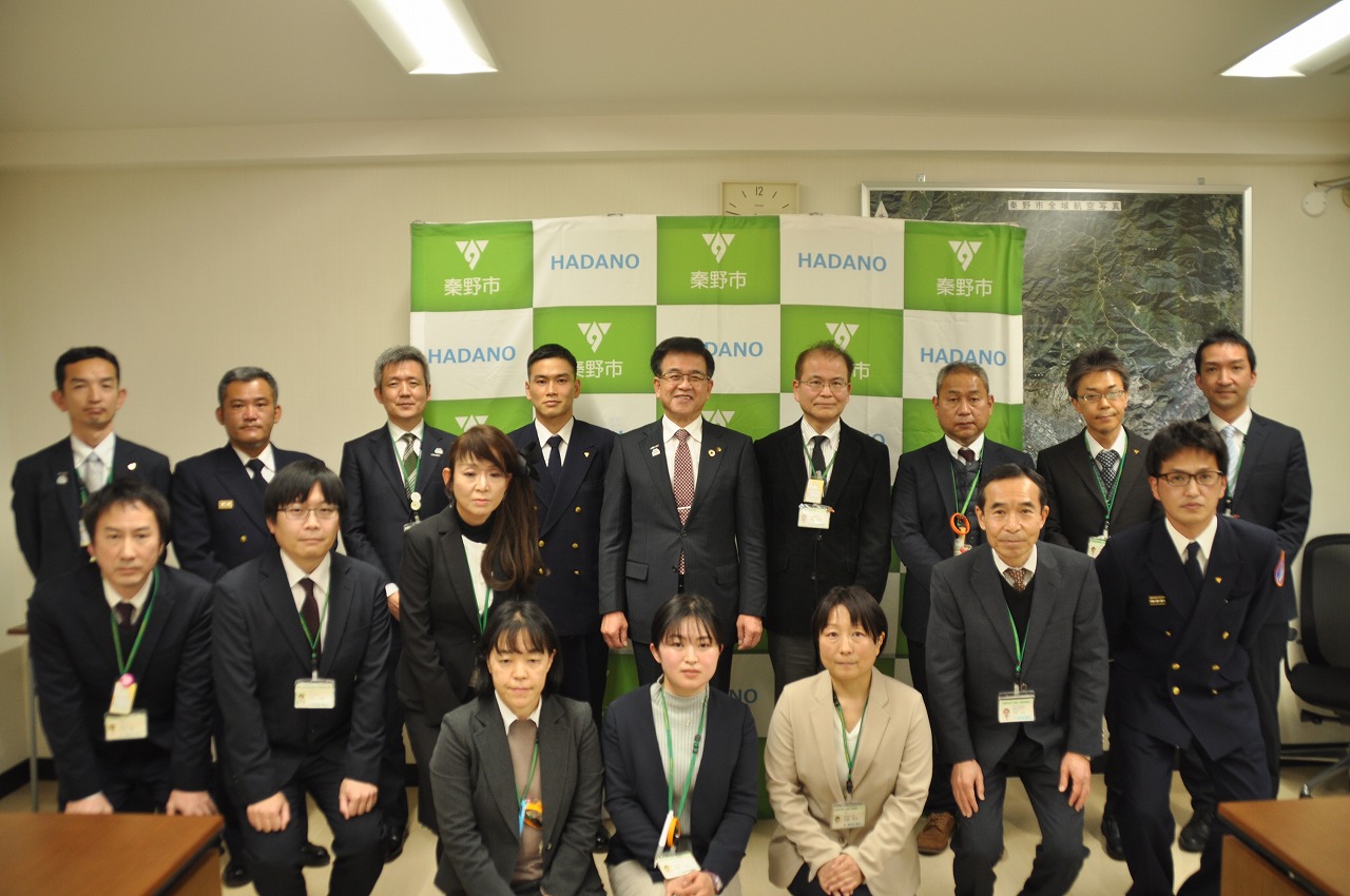 グリーンを基調とした市役所のロゴが並んだ背景の前で、スーツや制服姿の市長や複数の男女が立ちとしゃがみの二列になってカメラに向かい集合している様子を捉えた写真