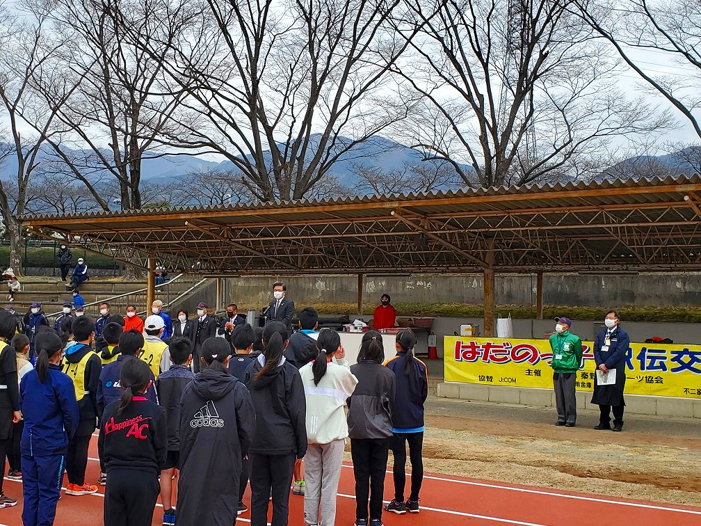 運動場の屋根付き休憩スペースの前に設置された演台に立った高橋市長が、トラックに整列した参加者の子どもたちに向かって話をしている様子の写真