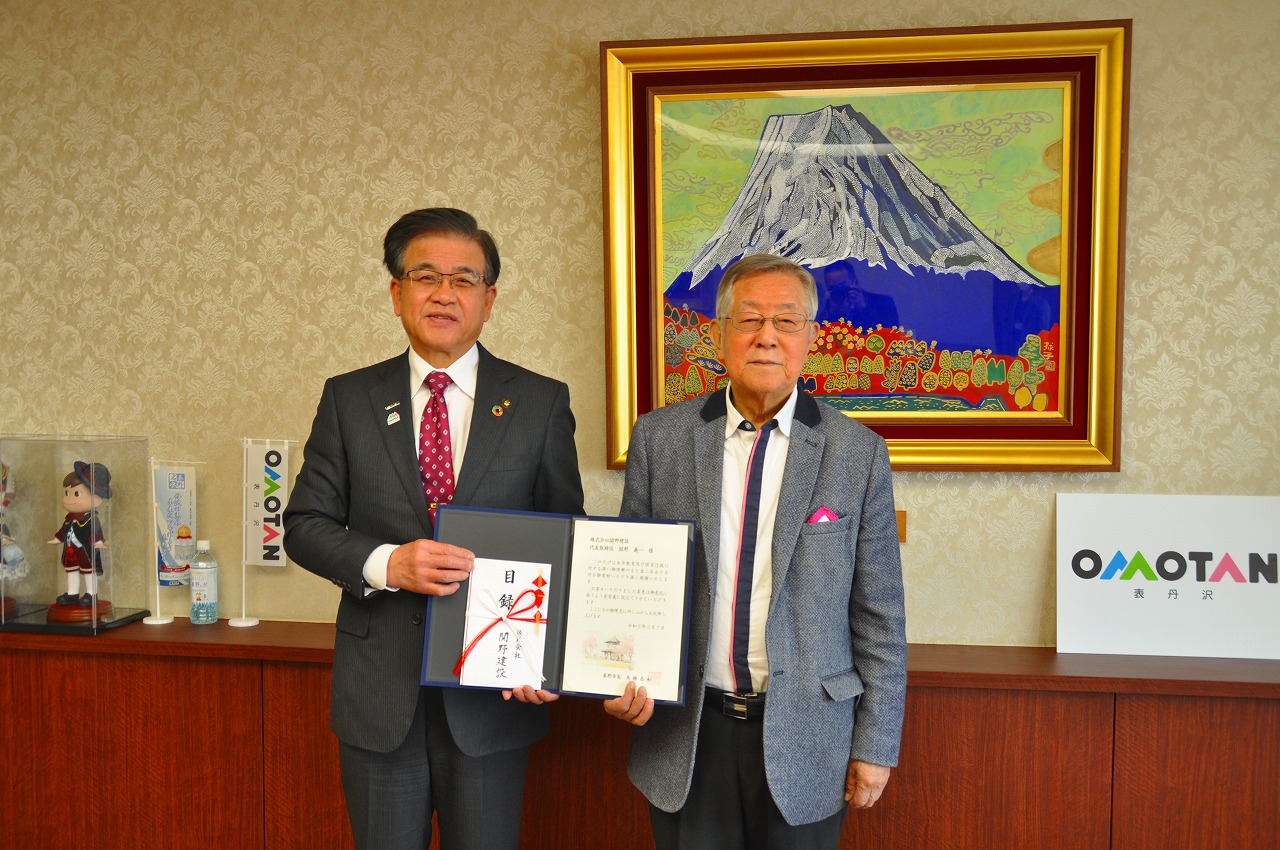 山の絵画が掛けられた壁を背に、高橋市長と株式会社関野建設・関野代表取締役が一緒に目録を持ち、並んで立っている記念写真