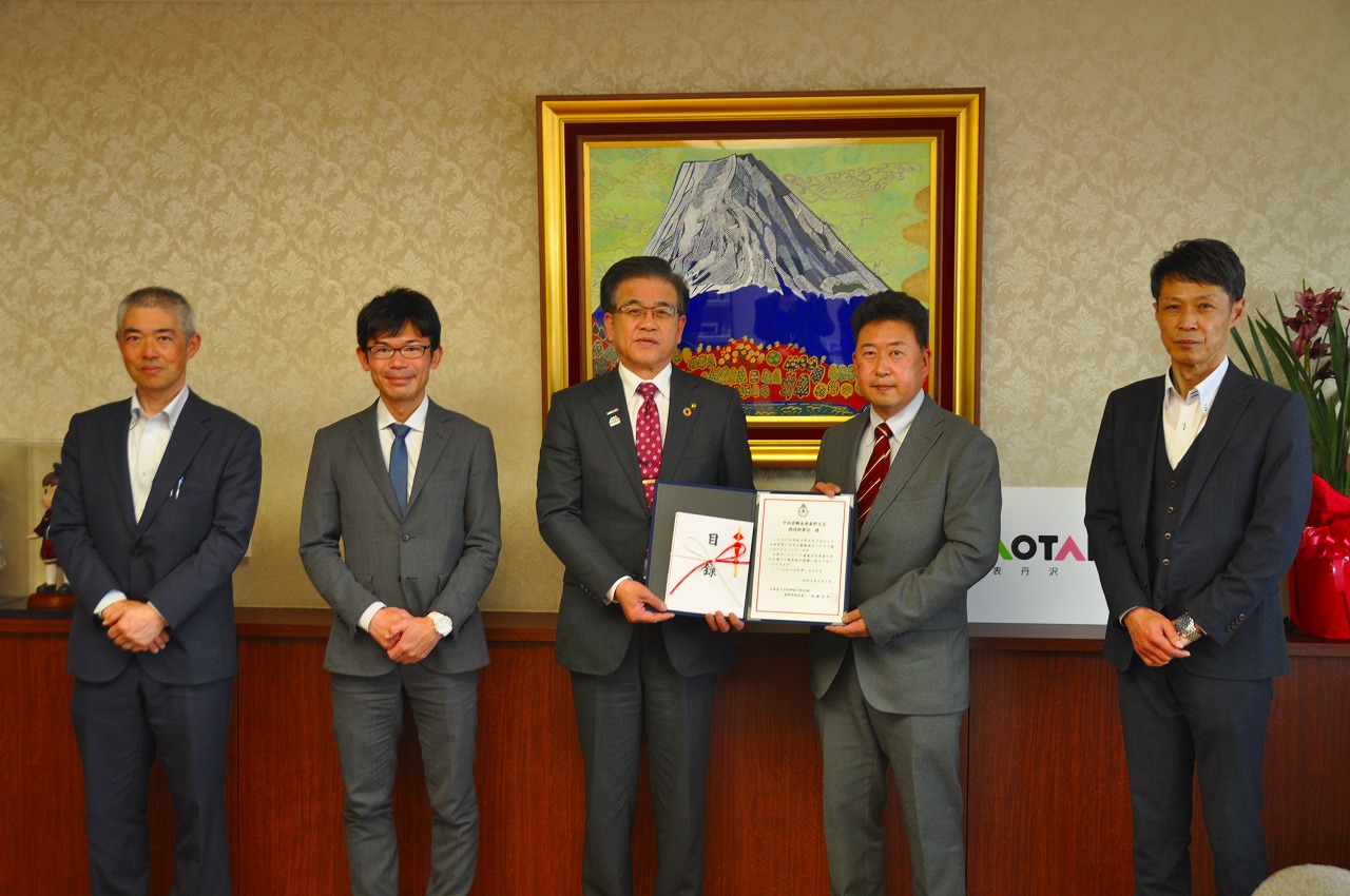 山の絵画が掛けられた壁を背に、高橋市長と関係者の男性が一緒に目録を持ち、他3人の男性と並んで立っている記念写真