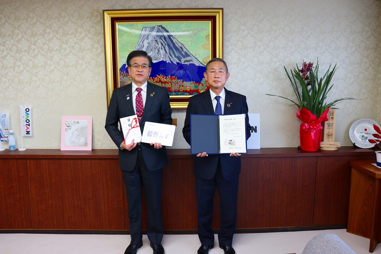 山の絵画が掛けられた壁を背に、高橋市長と平塚信用金庫・曽我専務理事がそれぞれ目録や証書を手に、並んで立っている記念写真
