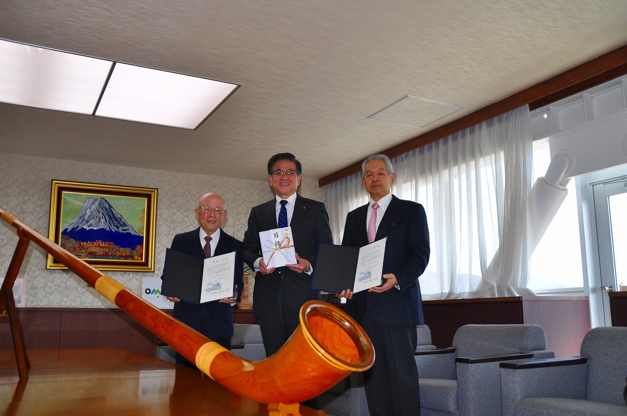 テーブルの上に置かれた大きなアルプホルンと、目録などを持ってその後ろに立った高橋市長、関係者の男性2人を写した記念写真