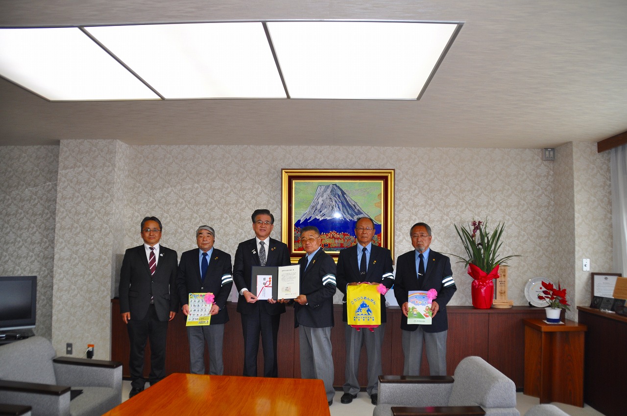 山の絵画が掛けられた壁を背に、目録を持った高橋市長と関係者の男性、パンフレットや袋を持った4人の関係者の男性が並んで立っている記念写真