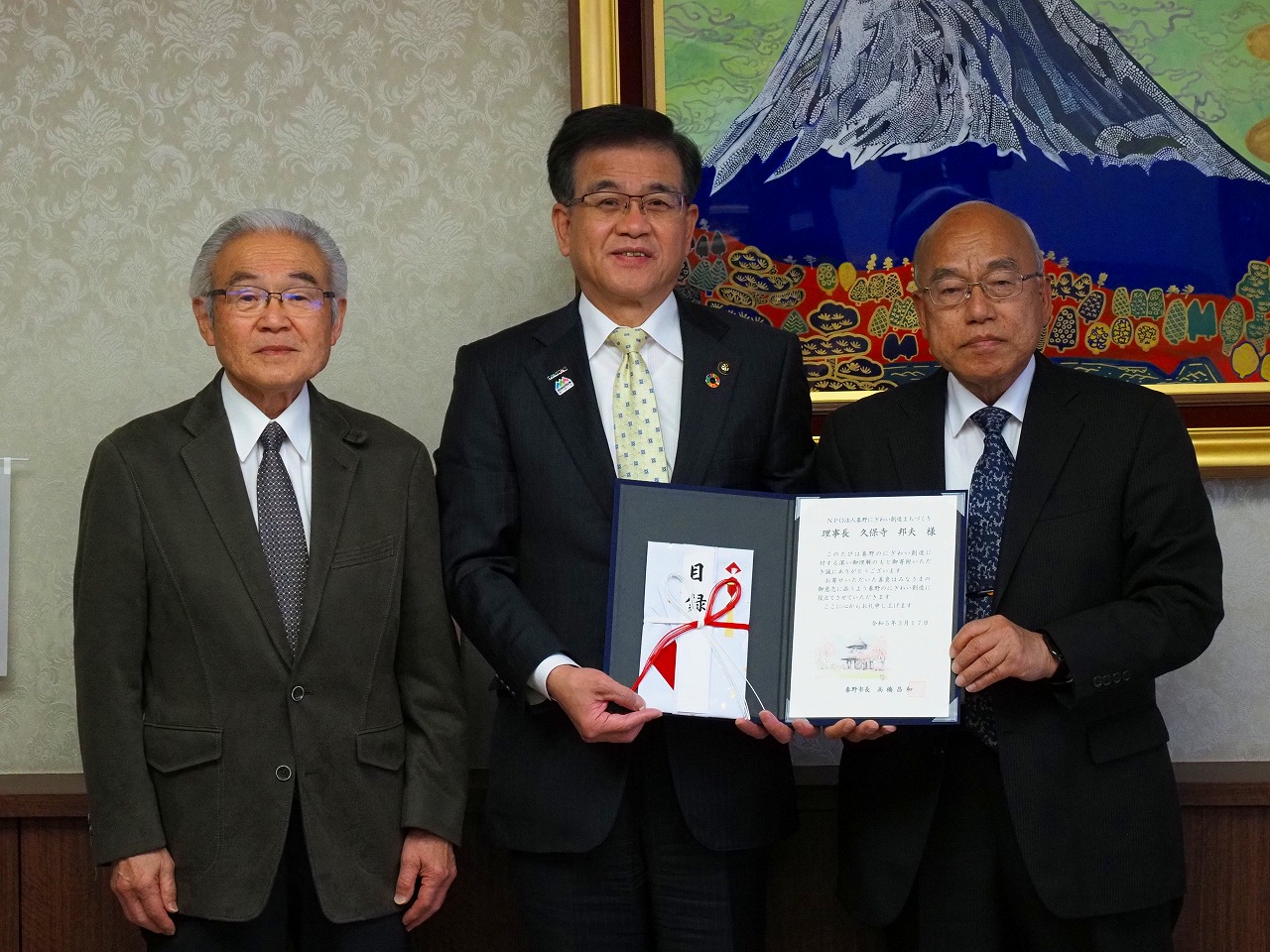山の絵画が掛けられた壁を背に、目録を一緒に持った高橋市長と関係者の男性、他1名の男性が並んで立っている記念写真