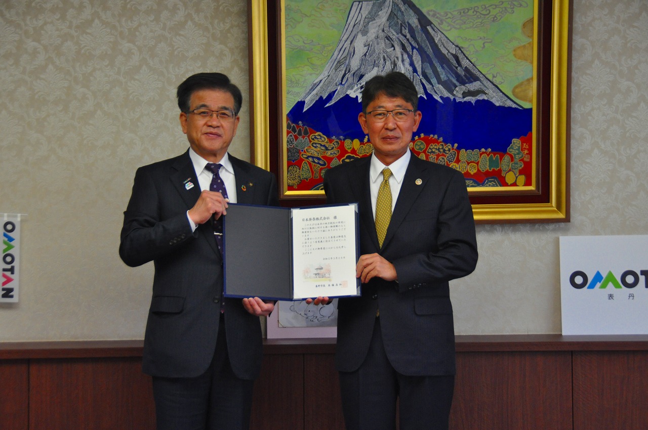 山の絵画が掛けられた壁を背に高橋市長と株式会社トープラ・新藤代表取締役社長が並んで立ち、一緒に証書を持って写した記念写真