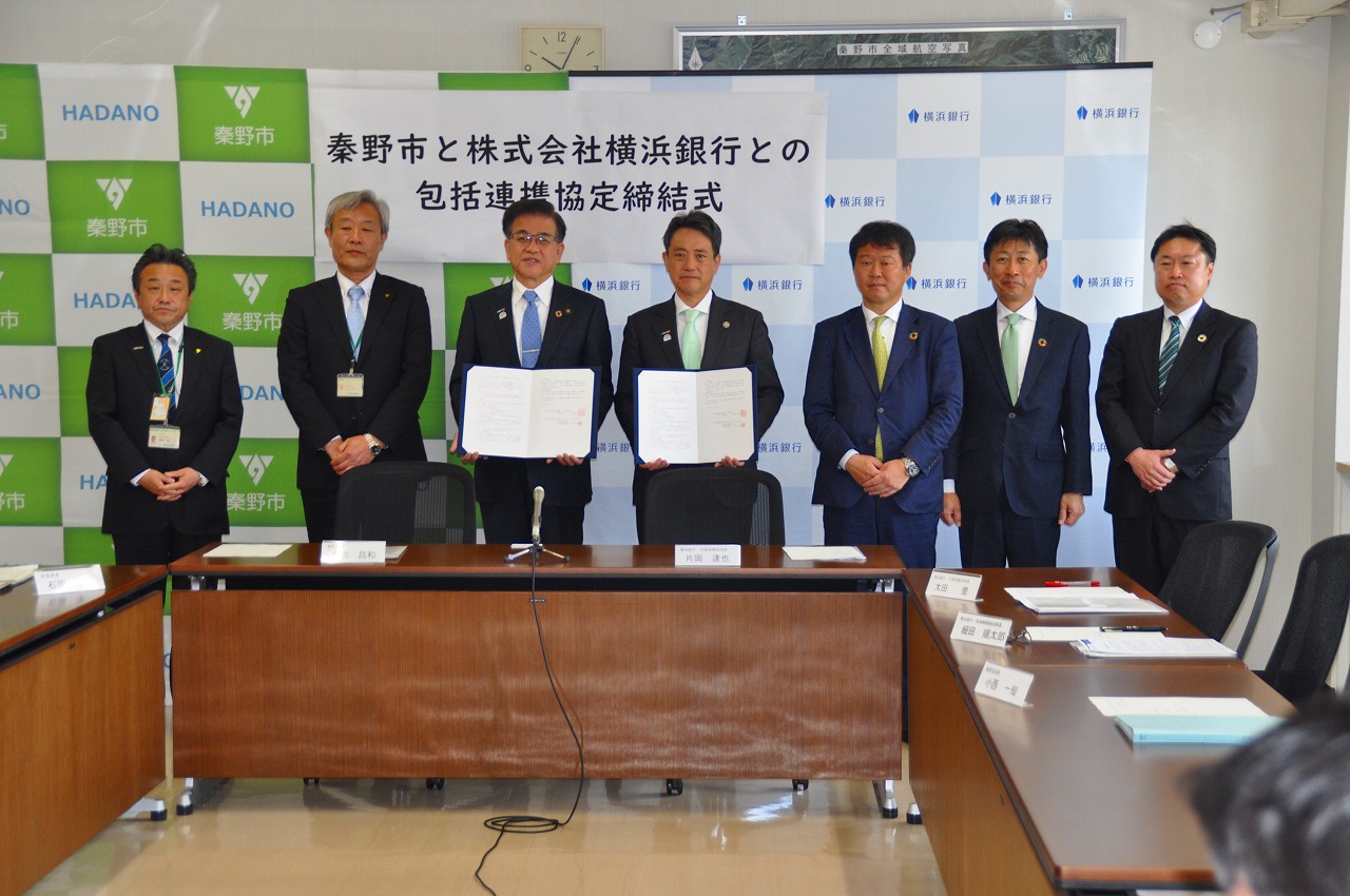 「秦野市と株式会社横浜銀行との包括連携協定締結式」の横断幕が掲げられたバックパネルの前で、中央で協定書を胸の前に持った高橋市長と関係者の男性を中心に、他関係者の男性が並んで立っている記念写真