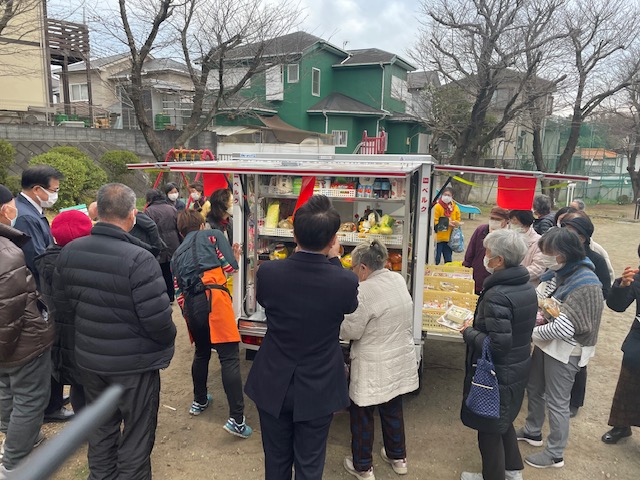 広場に停まった、たくさんの商品を積んだ移動式スーパーの車両の周りに大勢の買い物客が集まっている様子の写真