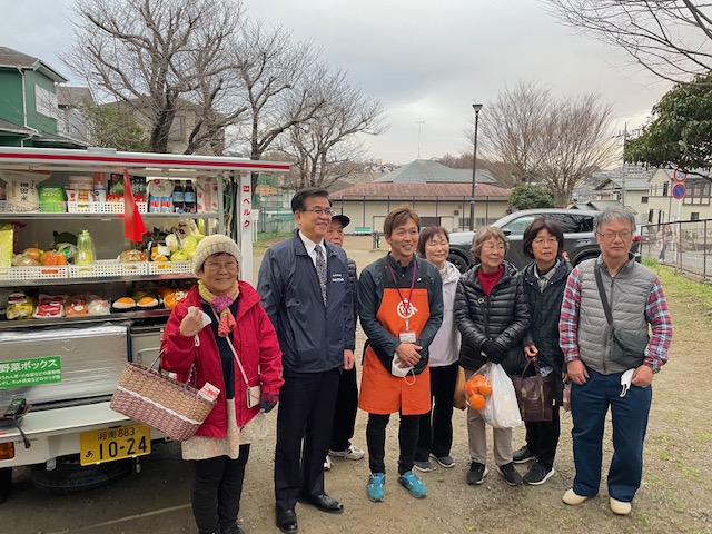 広場に停まった、移動式スーパーの車両にたくさんの野菜や果物、米などの食料品が積まれており、その前でカゴや袋を下げた数名の買い物客と、高橋市長、オレンジ色のエプロンをした男性が集まって記念撮影をしている写真