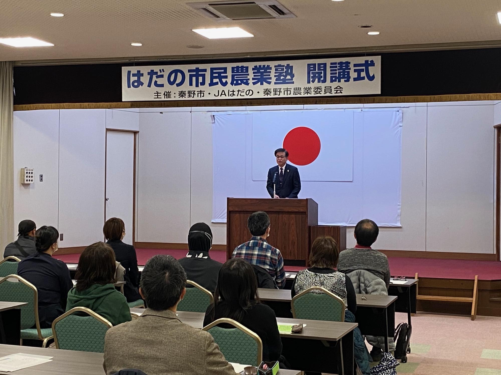 「はだの市民農業塾 開講式」の横断幕と日本国旗を背に、舞台の演台に立った高橋市長が席に座った複数の出席者たちに話をしている様子の写真