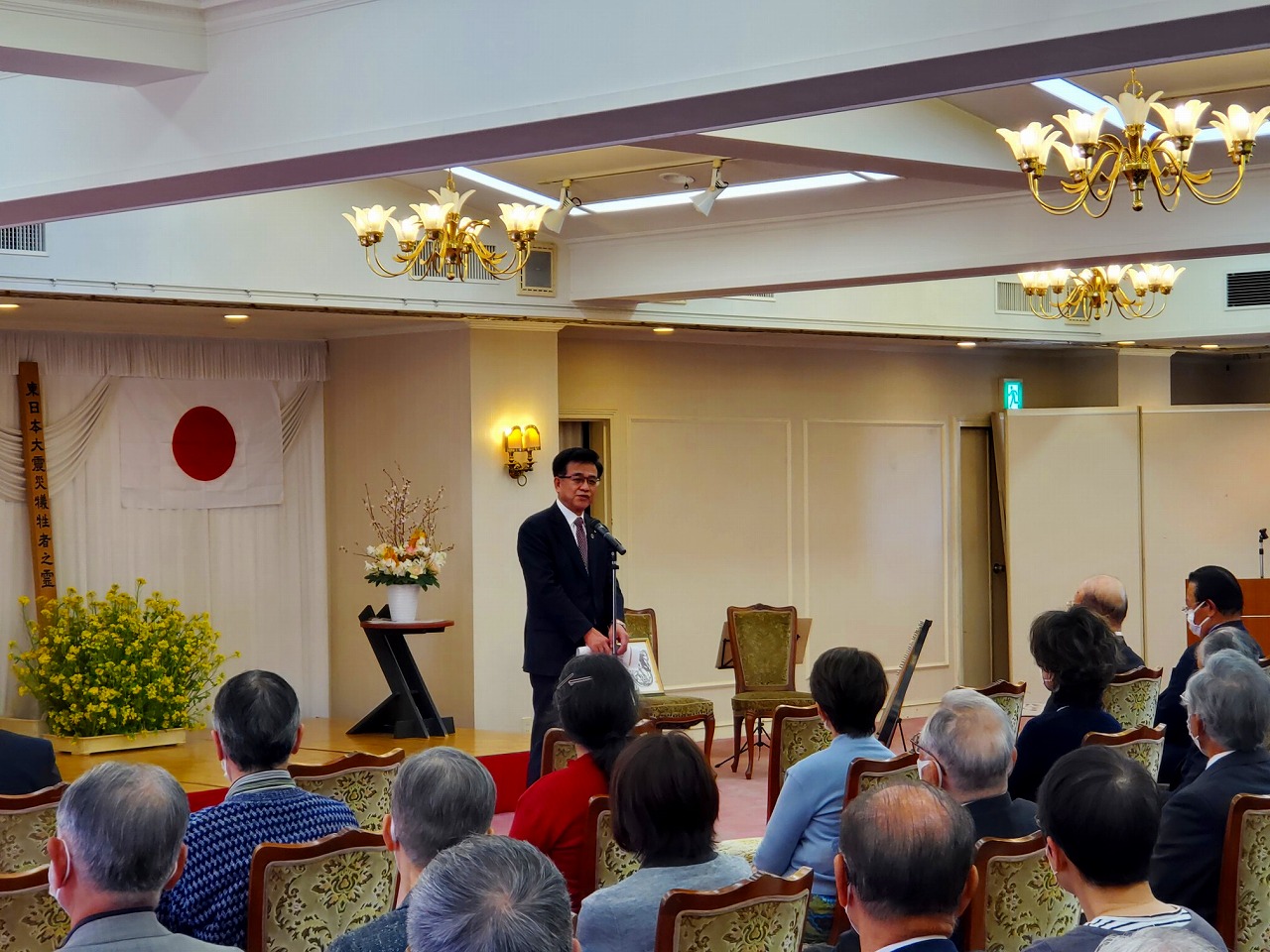 壁に日本国旗が掲げられた豪華なシャンデリアがある会場で、椅子に座ったたくさんの出席者に向かって、マイクの前に立った高橋市長が話をしている様子の写真
