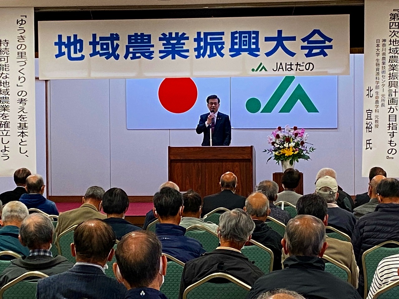 「地域農業振興大会 JAはだの」の横断幕が掲げられ、日本国旗とJAのマークが飾られた舞台の演台に立った高橋市長が、席に座った大勢の出席者に向かって話をしている様子の写真
