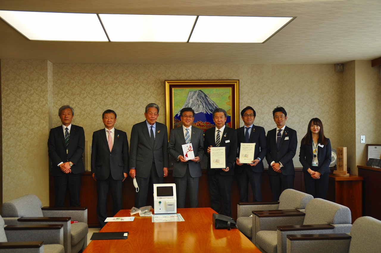 山の絵画が掛けられた壁を背に目録の封筒を持った高橋市長の左側に関係者の男性3人、右側に証書を持った2人の男性、関係者の男女が並んで立っている記念写真