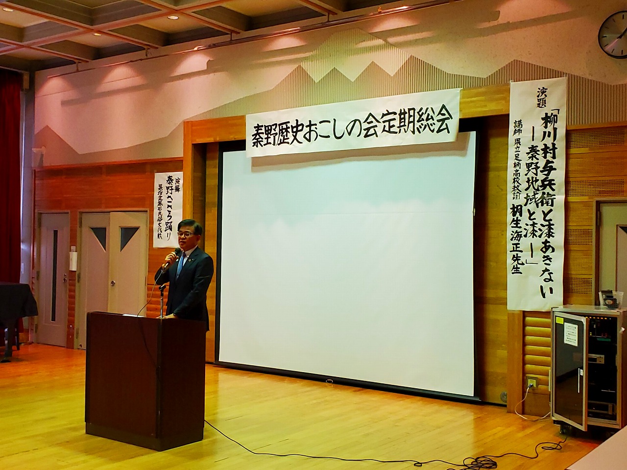 「秦野歴史おこしの会定期総会」と書かれた手書きの横断幕が大きなスクリーン上に貼られており、その前の演台に立った高橋市長が、マイクを持って話をしている写真