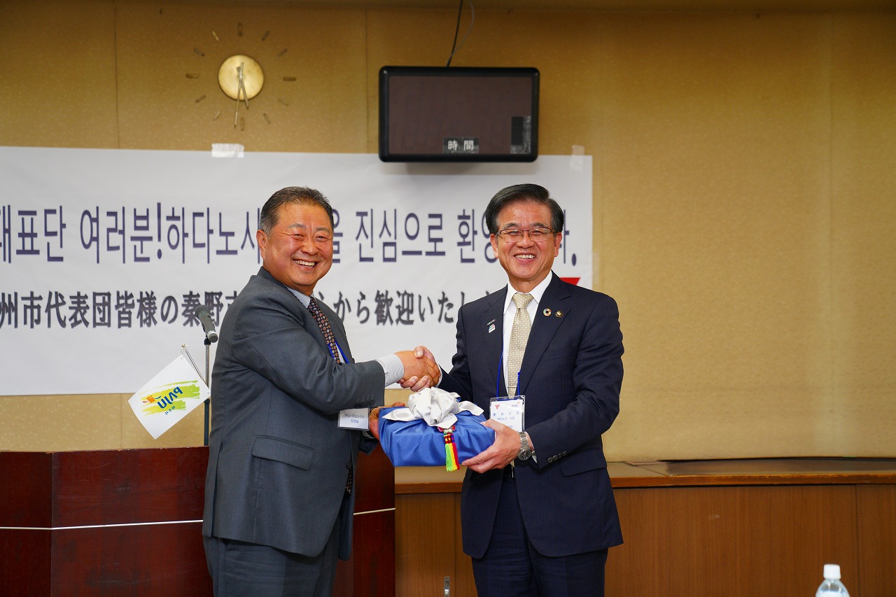韓国語と日本語で表記がされた歓迎メッセージが書かれた白い紙が貼られた壁の前で、片手に青い包みの箱を持った高橋市長が横に並んだ男性と握手をして笑顔で写っている記念写真