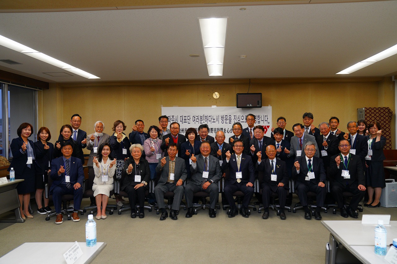 韓国語と日本語で表記がされた歓迎メッセージが書かれた白い紙が貼られた壁の前に、高橋市長と大勢の関係者たちが並び、前列の人々は椅子に座り、全員で親指と人差し指を交差させて指でハートを作るポーズをして笑顔で写っている記念写真