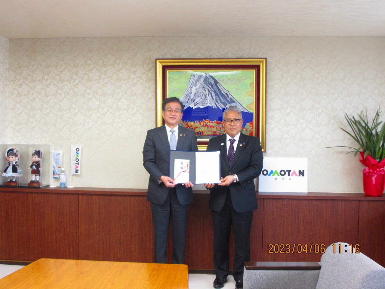 山の絵画が掛けられた壁の前で、高橋市長と中栄信用金庫・北村理事長が並んで立ち、一緒に目録を持って立っている記念写真