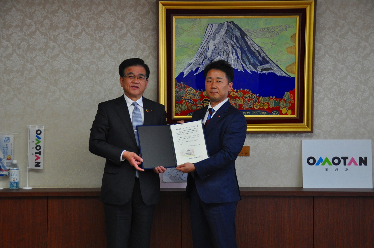山の絵画が掛けられた壁の前で、高橋市長と株式会社ティーズフューチャー・鈴木常務取締役が並んで立ち、一緒に証書を持って立っている記念写真