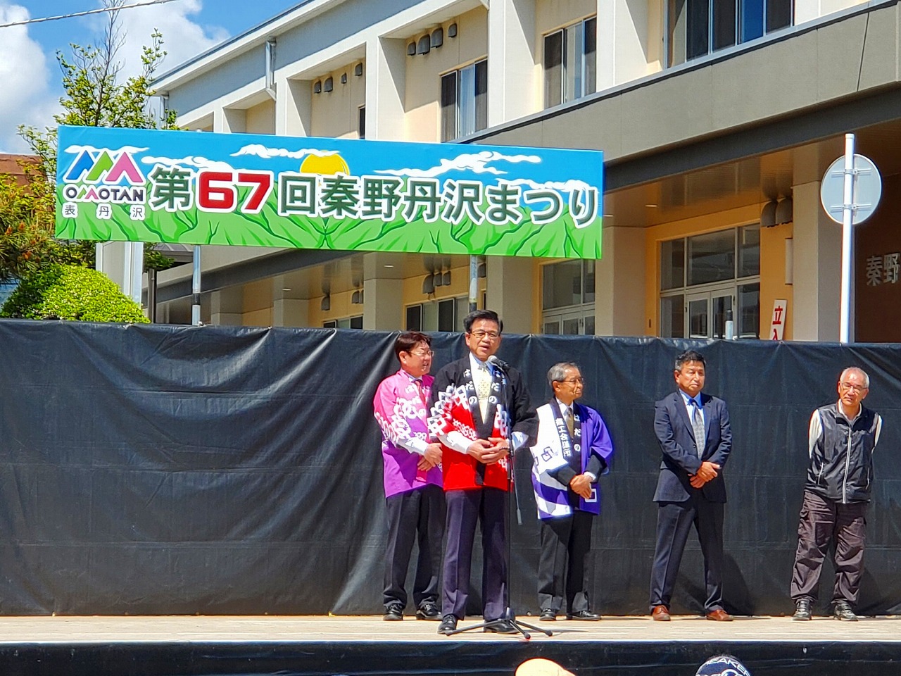 「第67回 秦野丹沢まつり」の横断幕が掲げられたステージの上に紫や青の法被を着た男性2名と、関係者の男性2人が立ち、その前に立った黒と赤の配色の法被を着た高橋市長がマイクに向かって話をしている様子の写真