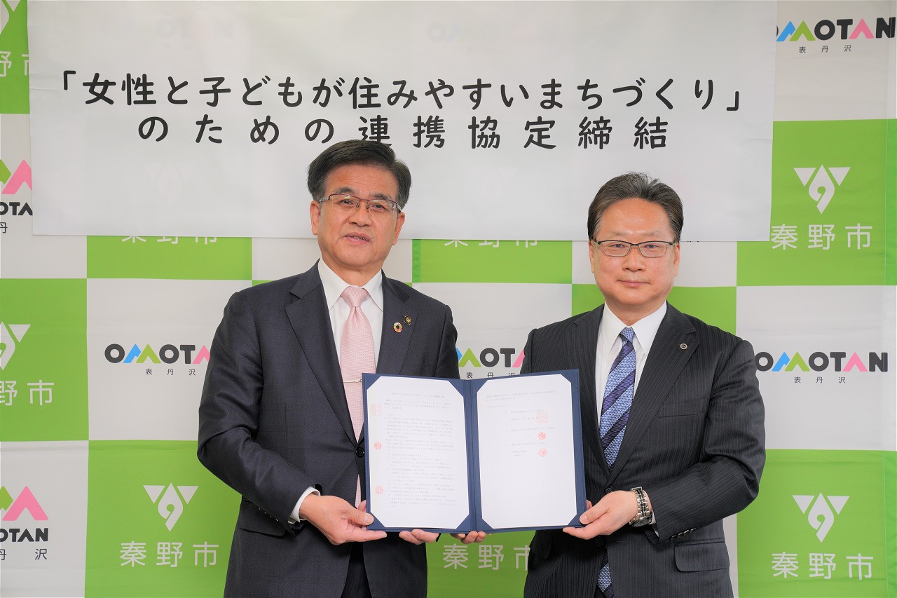 「「女性と子どもが住みやすいまちづくり」のための連携協定締結」の横断幕が貼られたバックパネルの前に、高橋市長と関係者の男性が並んで立ち、協定書を左右から持って写した記念写真