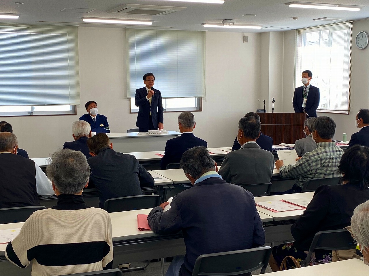 会議室に並んだ長机の席にたくさんの男性が座る中、前に立った高橋市長がマイクを持って話をし、その横には腕に腕章をつけた男性が着席し、右側の後ろに関係者の男性が立っている様子の写真