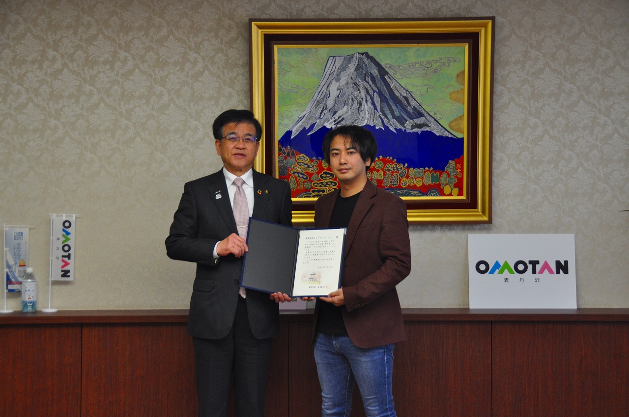 山の絵画が掛けられた壁の前で、高橋市長と株式会社リップルコミュニティ・山口代表取締役が並んで立ち、証書を持って写した記念写真