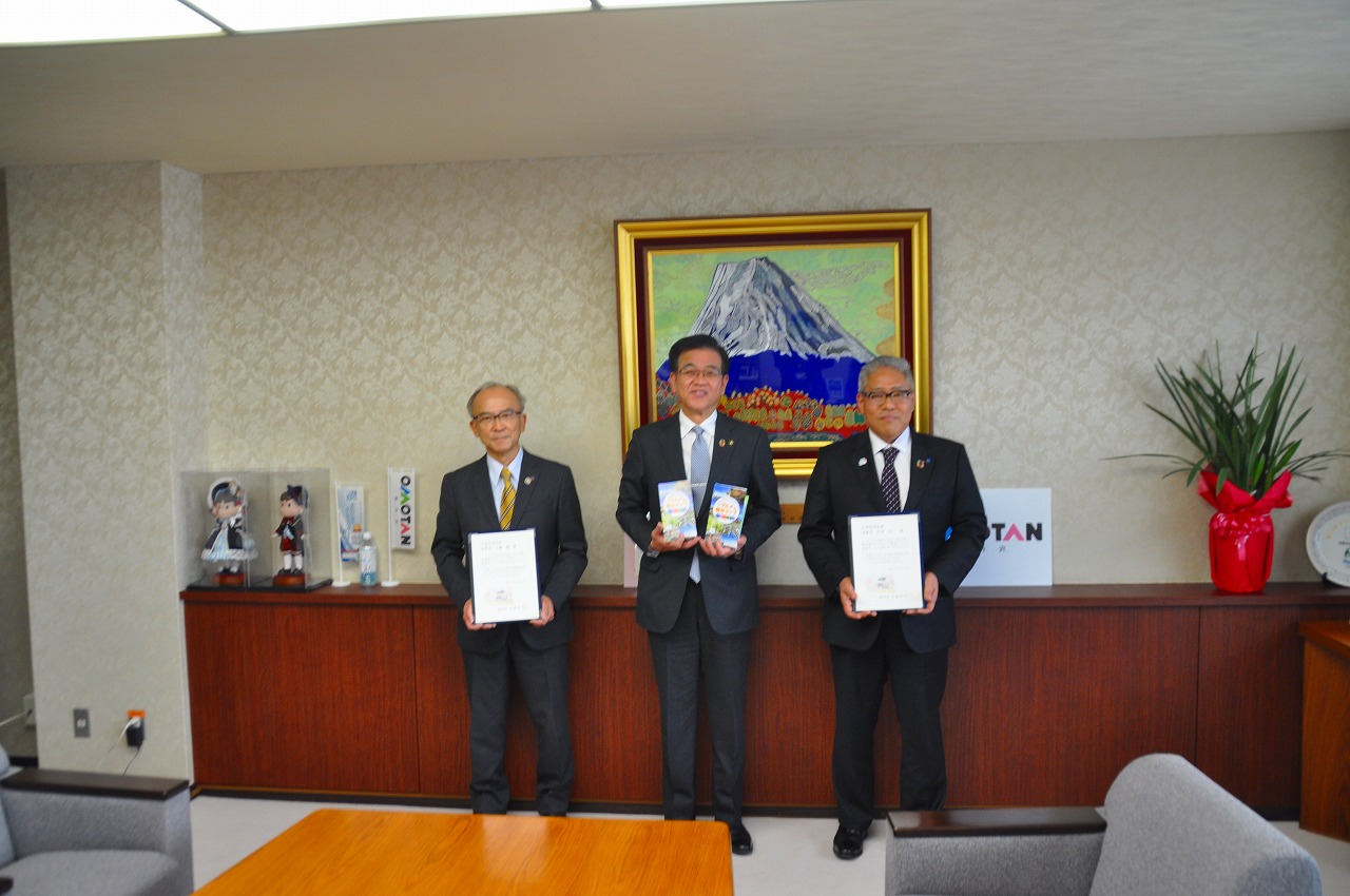 山の絵画が掛けられた壁の前で、両手に冊子を持った高橋市長の両隣に証書のようなものを持った中栄信用金庫・北村理事長と中南信用金庫・大藤理事長が並んで立っている記念写真