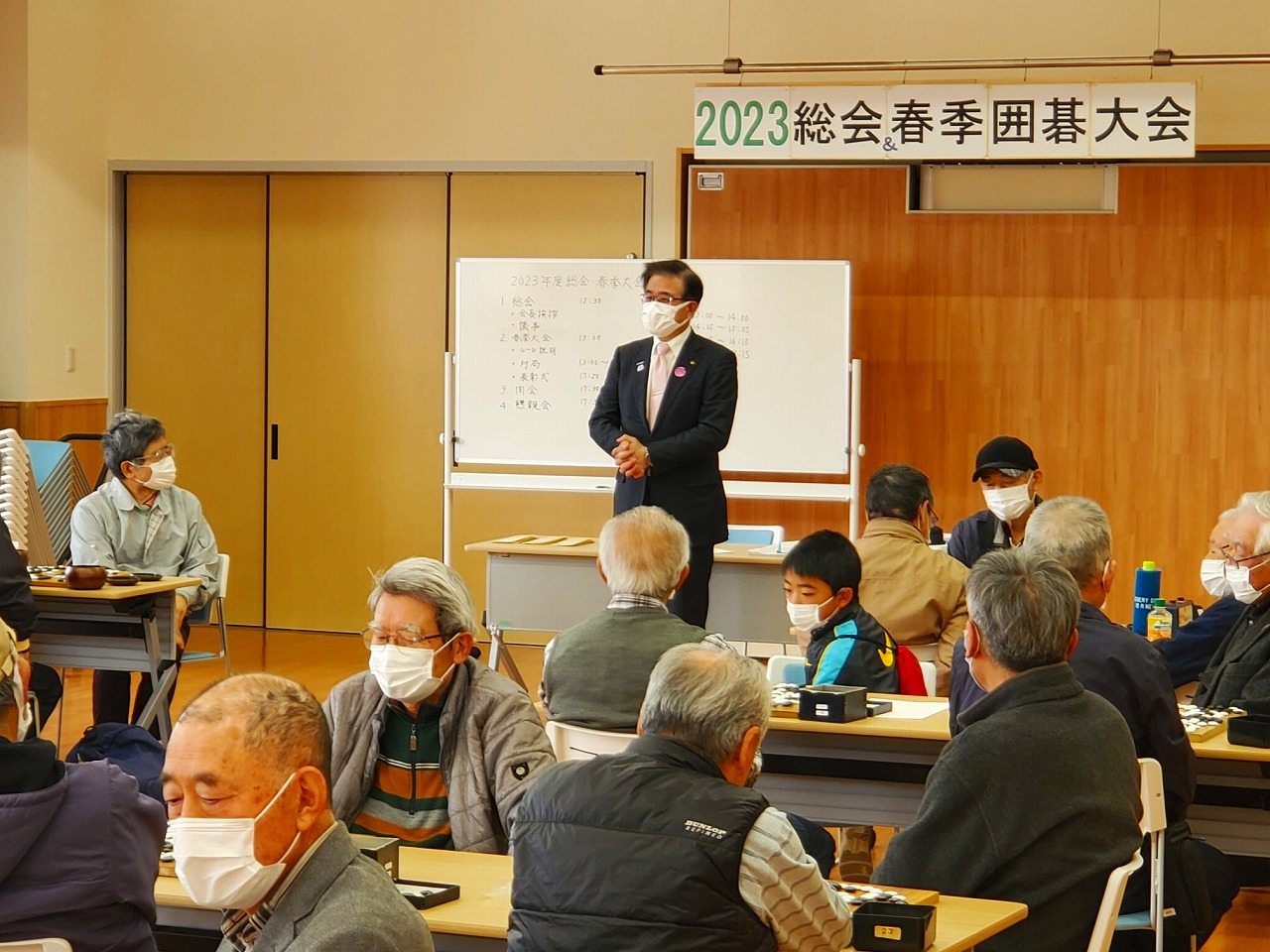 「2023総会春季囲碁大会」と書かれた横断幕の下で、ホワイトボードを背にして立った高橋市長が、複数並んだ碁盤が置かれたテーブルの席に座ったたくさんの参加者に向かって話をしている様子の写真