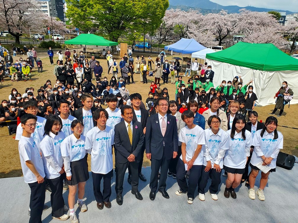 満開の桜の木々が見える芝生の広場に複数のテントが並び、大勢の人々が集まる中、ステージの上でおそろいの白いTシャツを着たたくさんの学生たちが高橋市長と関係者の男性を中央にして、広場を背景に記念写真を撮っている様子の写真
