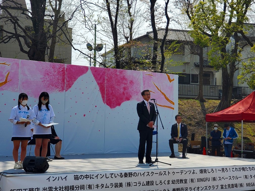 白地に淡いピンク色の桜の木々が描かれたパネルが設置されたステージの右側に白いTシャツを着た2人の女子学生が立ち、他の関係者数名が椅子に座る中、中央のマイクの前に立った高橋市長がスピーチをしている写真