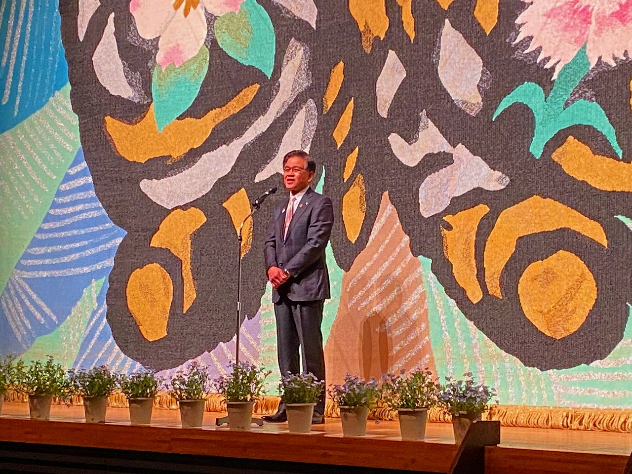 鉢植えの花が飾られ、背景に花などが描かれたステージの上で、スタンドマイクの前に立った高橋市長が話をしている様子の写真