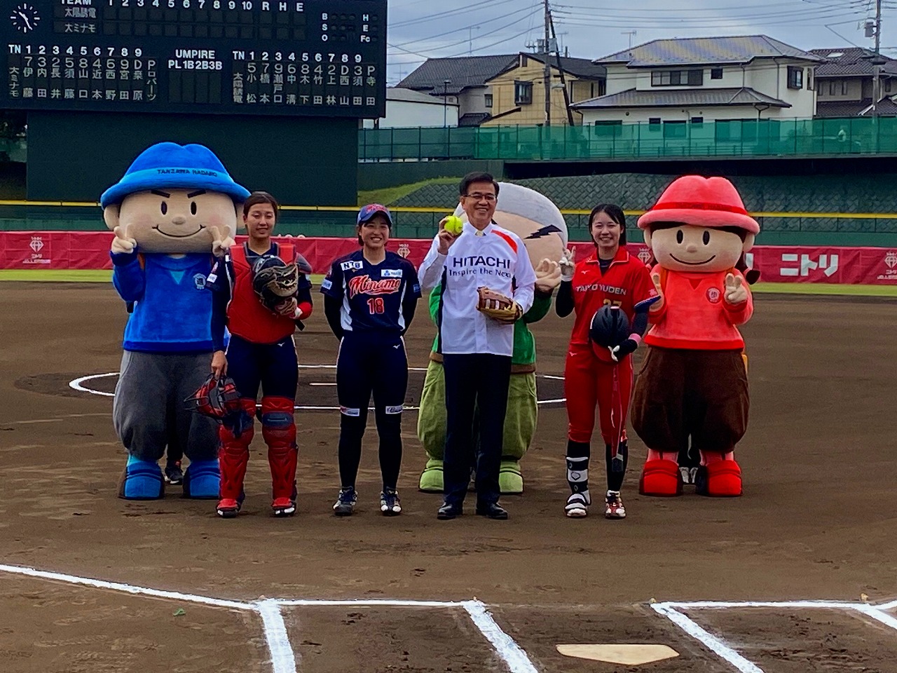 屋外球場で、丹沢はだの三兄弟のキャラクターとソフトボールのユニフォームを着てグローブなどを持った3人の女性選手の中央に立った高橋市長が、ソフトボールとグローブを手に笑顔で記念撮影している写真
