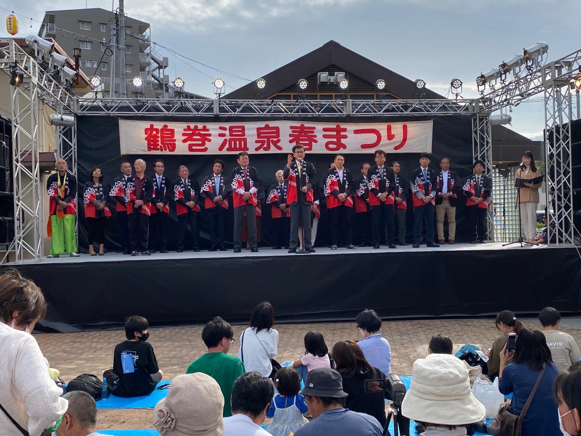 「鶴巻温泉春まつり」の横断幕が設置された屋外のステージの上に紺色と赤の配色の法被を着たたくさんの関係者が並んで立ち、手前に立った高橋市長がステージ下に集まった人々に向かって話をしている様子の写真