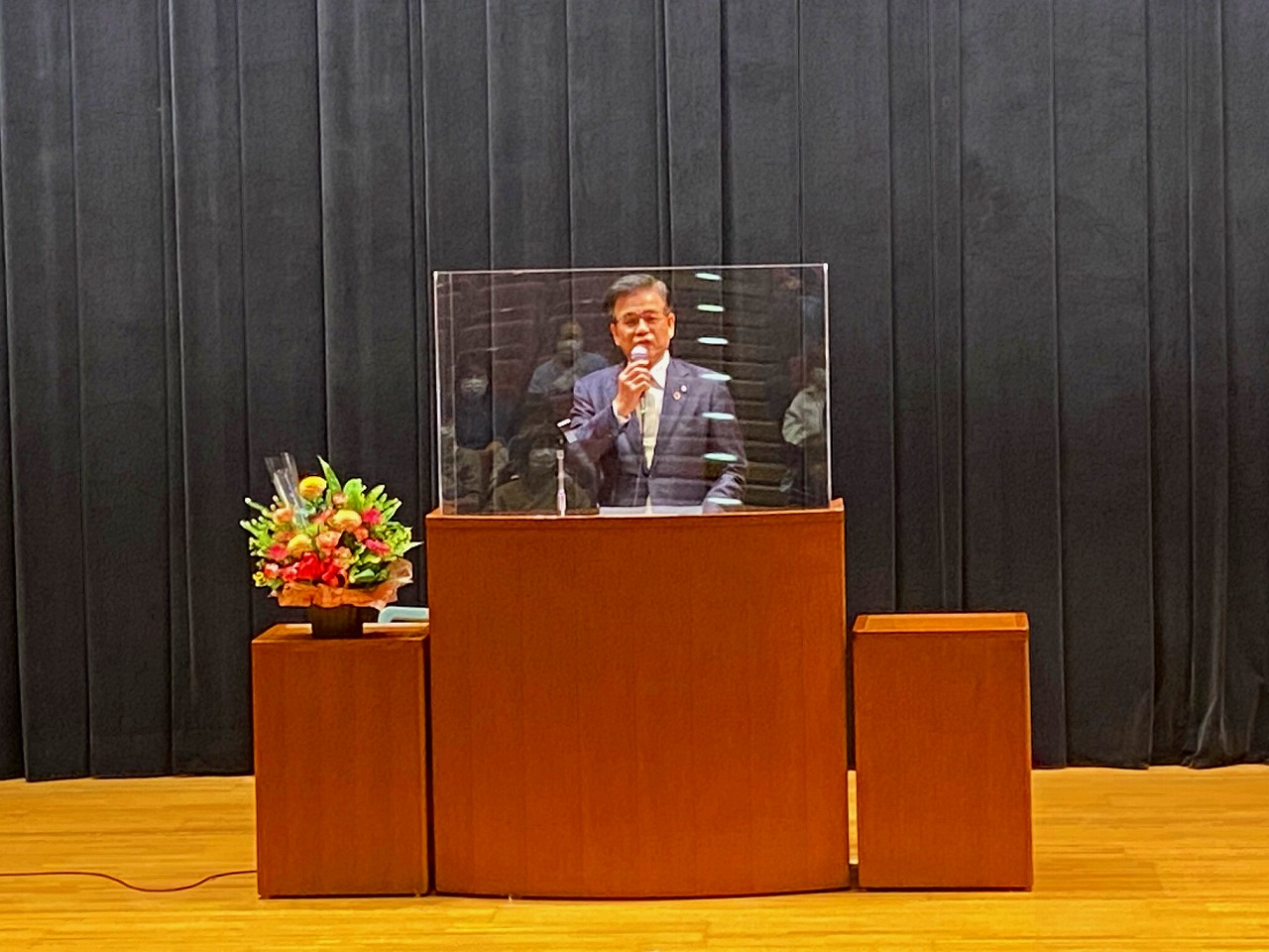 花が飾られ、アクリルパネルが設置された演台に立った高橋市長が、マイクを持って話をしている写真