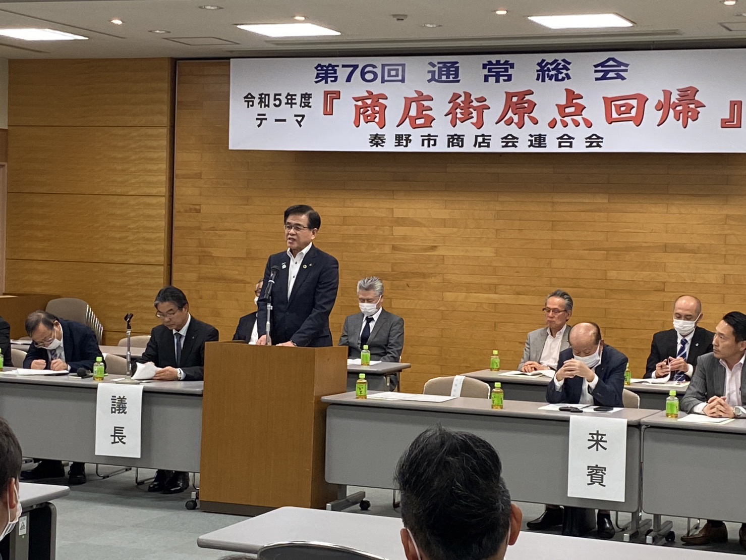 秦野市内のホールで「第76回通常総会」と書かれた横断幕の下、壇上に立つ市長が挨拶をしており、議員や議長、来賓の方々が机に座って話を聞いている様子の写真