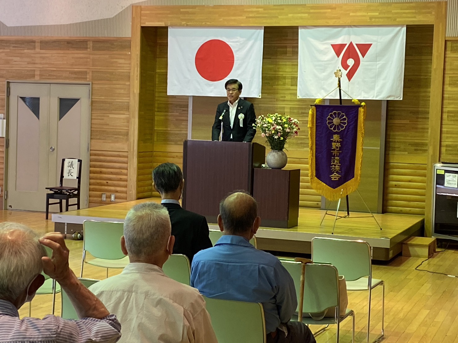 日の丸の旗と秦野市遺族会の紫色の旗が飾られた壇上で、市長が演説をしており、その様子を複数の人々が椅子に座って聞いている様子の写真