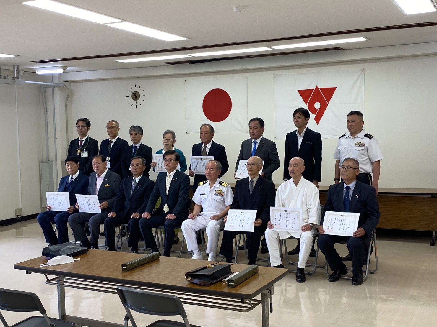 白を基調とした部屋で、日の丸の旗と市旗を背景に、スーツや制服姿の男性たちが委嘱状のようなものを持って2列に並んでいる集合写真