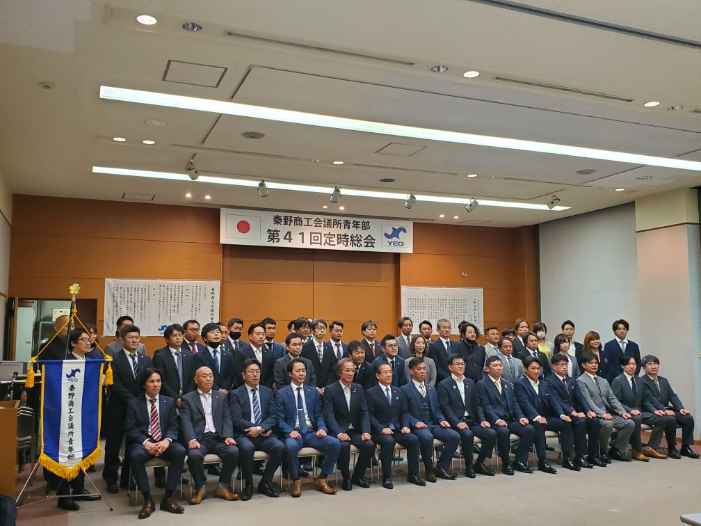 「秦野商工会議所青年部第41回定時総会」と書かれた看板の下で、スーツを着たの複数の男性と数人の女性が集まって並んでいる集合写真
