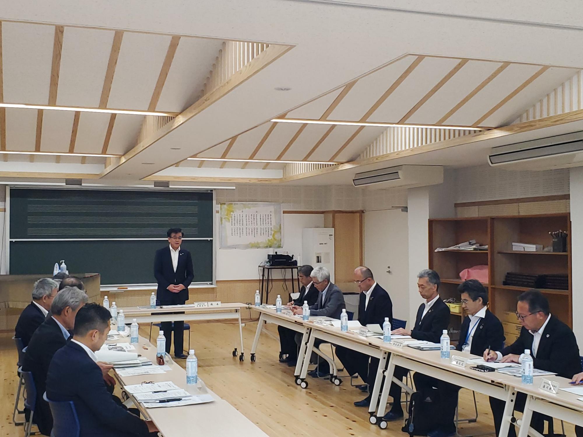 会議室で、スーツ姿の市長が中央に立ち、それを囲むように座っている複数の人々が真剣な表情で会議に参加している様子の写真