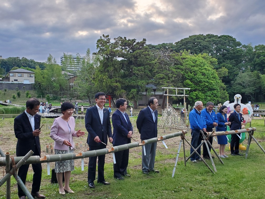 複数の人たちが屋外に設置された竹の灯篭の前に並んでおり、手前にいる男性2人と女性1人が笑顔でこちらを見て、奥の人たちは真剣な表情をしている様子の点灯式での写真