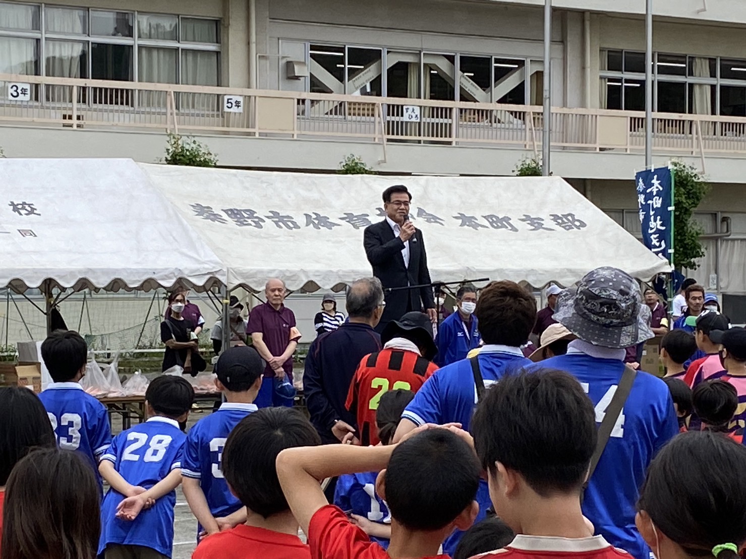 グラウンドのテントの前で、市長がマイクを持ってスピーチをしており、たくさんの子供たちと大人たちがその話を聞いている様子の写真