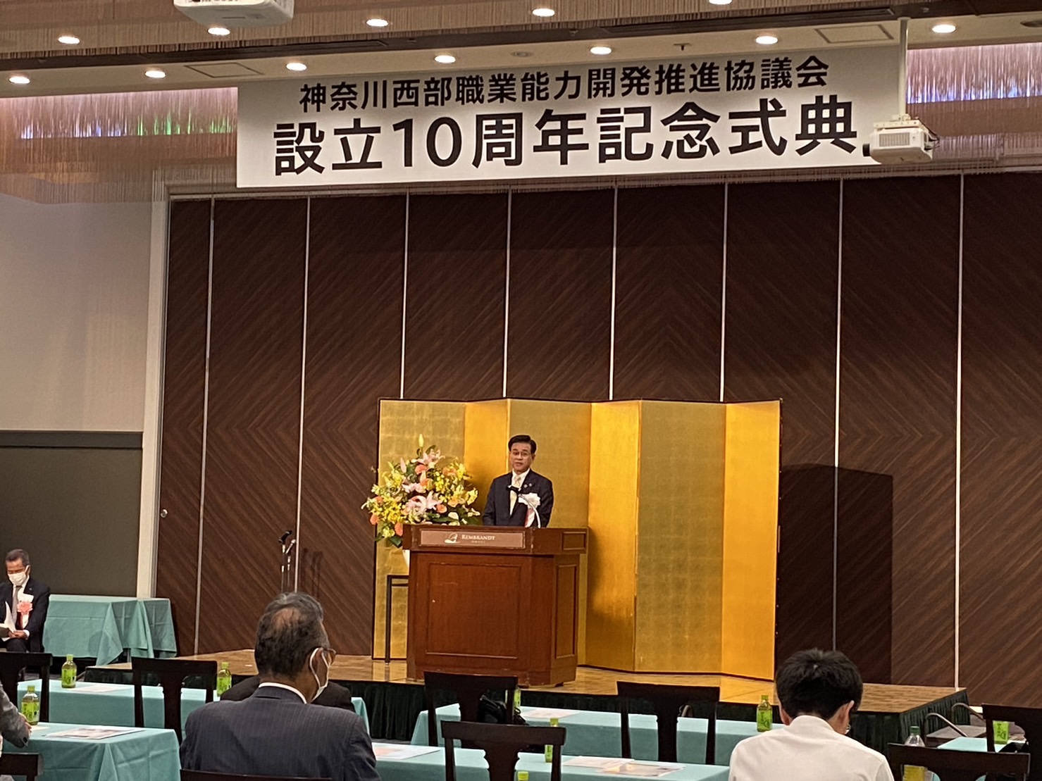 「神奈川西部職業能力開発推進協議会設立10周年記念式典」と書かれた横断幕が掲げられ、茶色の壁と金屏風を背景にした壇上で、市長が演説をしている写真