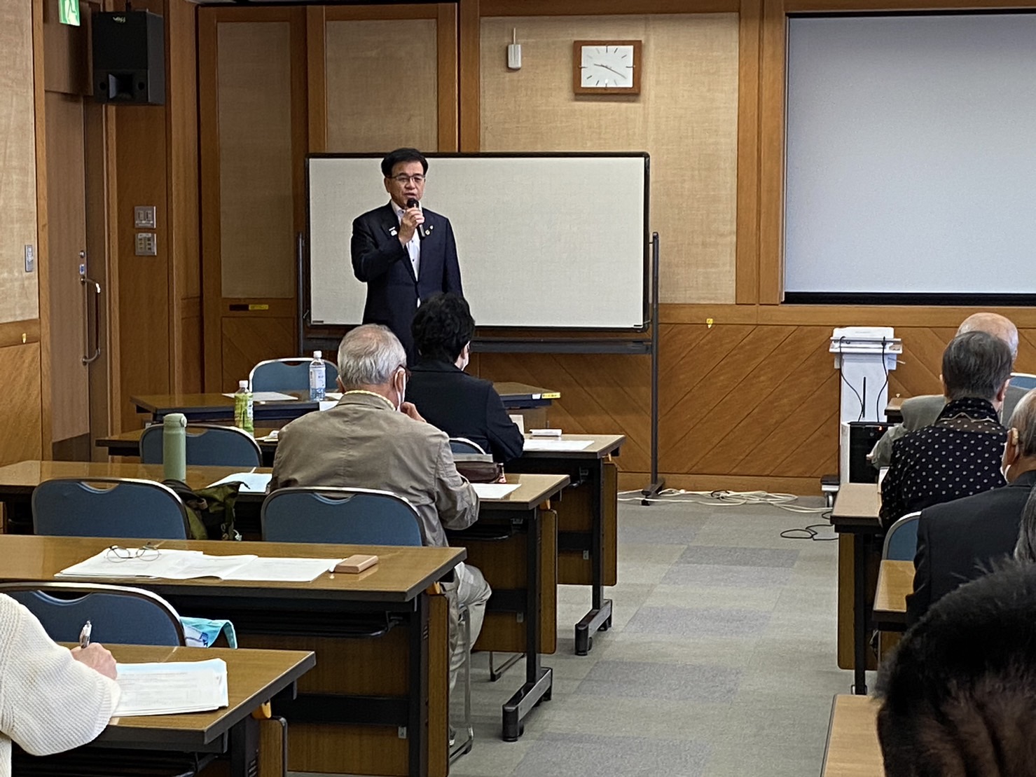 ホワイトボードの前で1市長が聴衆に向かってマイクを持ってスピーチをしており、出席者たちが整然と並んだ机と椅子に座って聞いている様子の写真