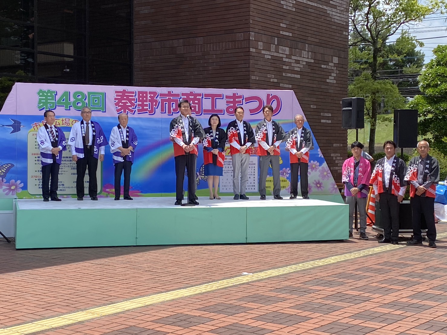 「第48回秦野市商工まつり」の看板があるステージ上で、お揃いの黒と赤の法被を着た4名の男性と一人の女性、その斜め後方に白と紫のお揃いの法被を着た男性3名が開会式に参加している様子の写真