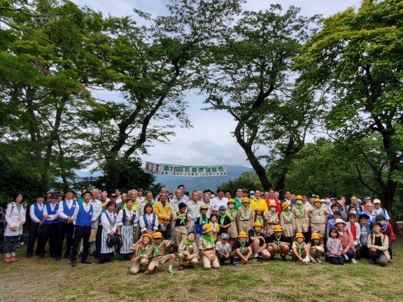 たくさんの老若男女が木の生い茂った野外で、子供たちはスカウトの制服、大人たちはそれぞれ様々な服装で集まって並んでいる集合写真