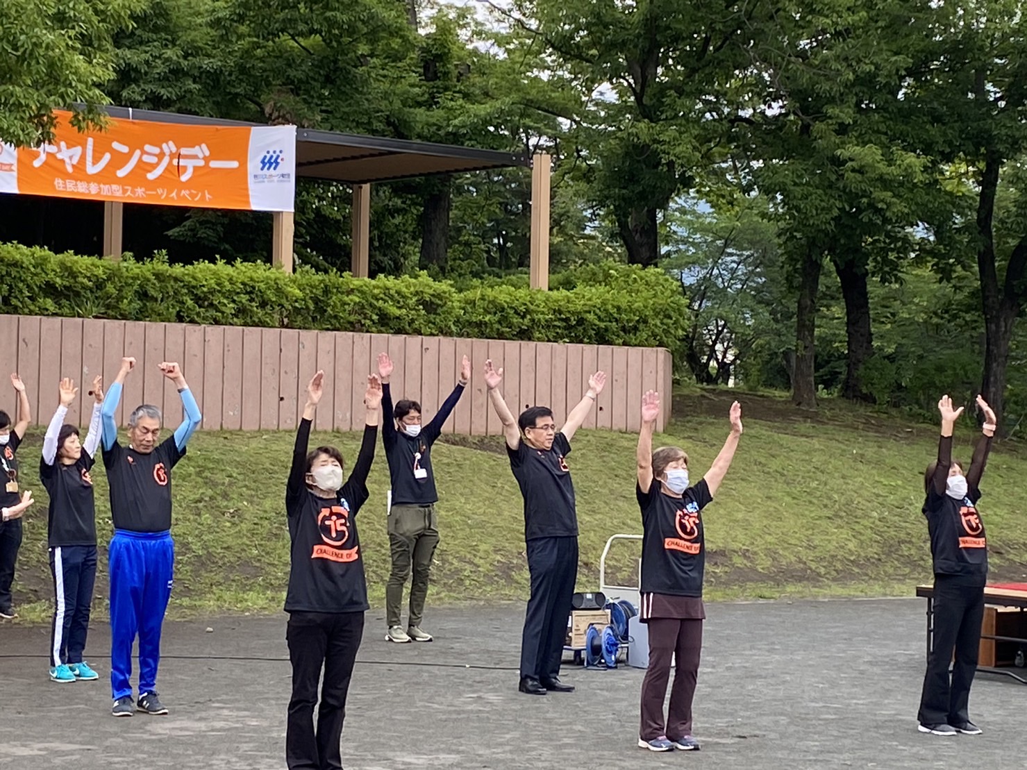 緑豊かな公園の広場で、年齢も性別も様々な人々が、黒いTシャツを着て両手を挙げ、体操をしている写真